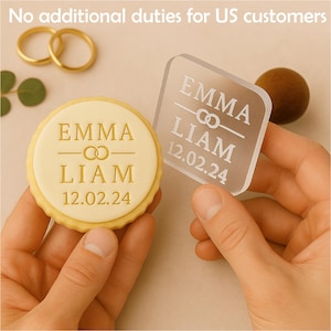 May include: A round cookie and a clear square acrylic piece, both with the names "EMMA" and "LIAM" and the date "12.02.24" engraved. The cookie has a yellow border and a cream-colored center. Two gold rings are in the background.