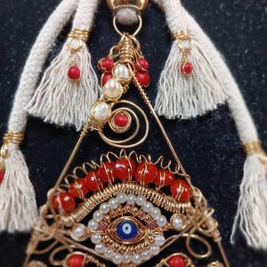 May include: A gold wire wrapped triangle pendant with a blue and white evil eye charm, surrounded by red and white beads. The pendant is attached to a white rope with tassels.