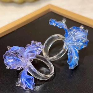Puede incluir: Dos servilleteros de cristal transparente con detalles de flores azules y moradas. Los pétalos de las flores tienen un aspecto delicado y translúcido. Los anillos están sobre una superficie negra.