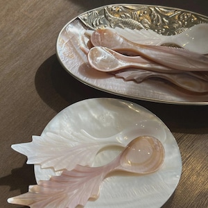 May include: A collection of mother-of-pearl spoons and serving dishes. The spoons have a leaf-like design and a pearlescent pink hue. The serving dishes are oval and round, with a silver trim and a pearlescent finish.