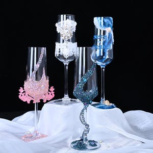 Puede incluir: Cuatro copas de champán con adornos decorativos. Una está adornada con purpurina rosa y una base rosa, otra con encaje blanco y una tercera con cinta azul. La cuarta copa tiene purpurina azul y un tallo de cuentas.