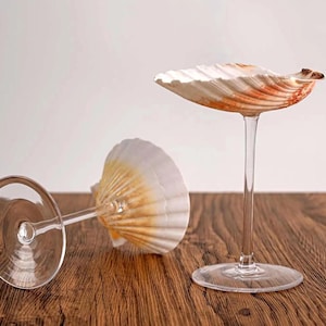Peut inclure: Deux verres à vin décoratifs avec des coquillages en guise de coupes. L'un est debout, l'autre est couché sur le côté. Les coquillages sont blancs avec des accents orange et marron. Les verres sont posés sur une surface en bois.