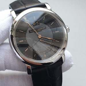 Könnte beinhalten: Eine automatische Armbanduhr von Baume & Mercier mit schwarzem Lederarmband. Die Uhr hat ein silberfarbenes Gehäuse, ein dunkelgraues Zifferblatt mit goldfarbenen Zeigern und Markierungen und das Datum bei 12 Uhr.