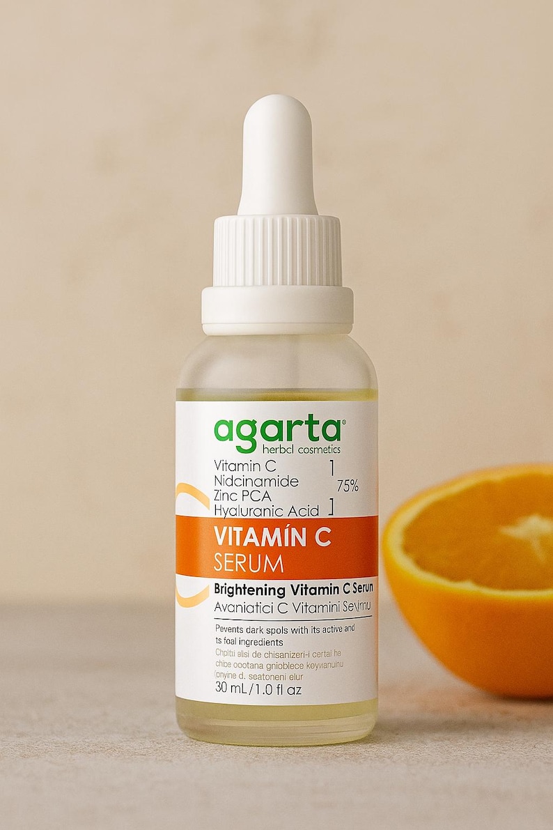 Op de afbeelding: Een heldere glazen fles vitamine C serum met een witte druppelaar, oranje label en de tekst "agarta herbal cosmetics". Het serum is gelabeld "VITAM&Iacute;N C SERUM" en bevat een sinaasappelschijf.