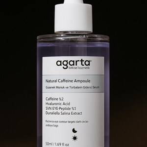 Op de afbeelding: Een heldere glazen fles Agarta Natural Caffeine Ampoule met een witte druppelkop. Het etiket bevat tekst over de ingrediënten en voordelen van het product, waaronder cafeïne 2% en hyaluronzuur. De fles bevat 50 ml.