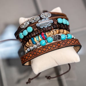 Op de afbeelding: Een stapel armbanden in bohemien-stijl. De armbanden hebben bruin leer, turquoise kralen, houten kralen en zilverkleurige metalen accenten. Eén armband heeft een geometrisch patroon. De armbanden worden gepresenteerd op een crèmekleurig kussen.