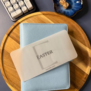 May include: A light blue textured binder with a translucent overlay card featuring the word EASTER and the text KIMMYPBUDGETS. The binder is placed on a round wooden tray next to a white calculator, a small blue ceramic dish, and two dice.