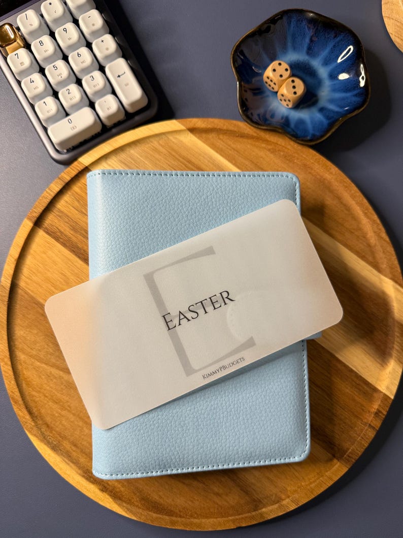 May include: A light blue textured binder with a translucent overlay card featuring the word EASTER and the text KIMMYPBUDGETS. The binder is placed on a round wooden tray next to a white calculator, a small blue ceramic dish, and two dice.
