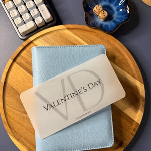 May include: A light blue textured binder sits on a round wooden tray. A white rectangular card with the text VALENTINE'S DAY and KIMMYPBUDGETS rests on top. Nearby are a white calculator and a small blue ceramic dish containing two wooden dice.