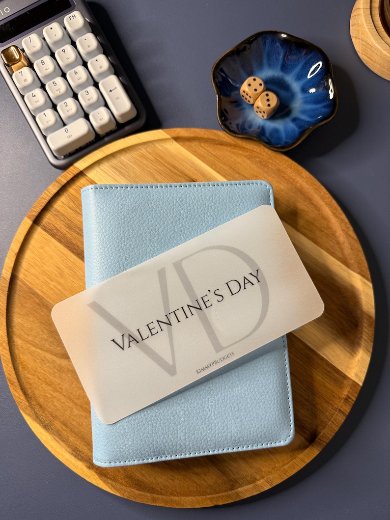 May include: A light blue textured binder sits on a round wooden tray. A white rectangular card with the text VALENTINE'S DAY and KIMMYPBUDGETS rests on top. Nearby are a white calculator and a small blue ceramic dish containing two wooden dice.