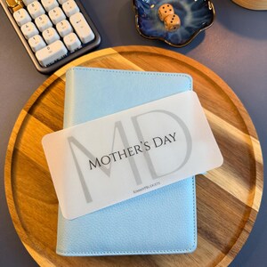 May include: A light blue textured binder with a white rectangular card placed on top. The card features the text MOTHER'S DAY in black lettering with a large grey MD background and the small text KIMMYPBUDGETS at the bottom center.