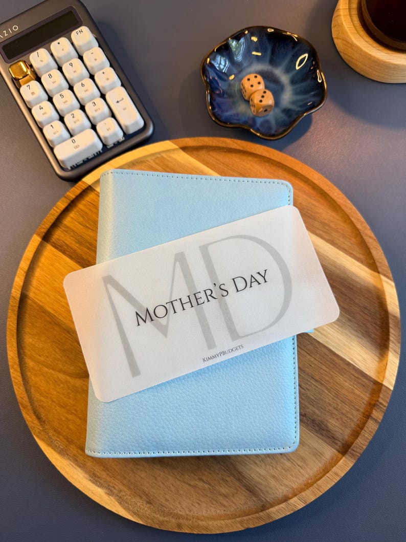 May include: A light blue textured binder with a white rectangular card placed on top. The card features the text MOTHER'S DAY in black lettering with a large grey MD background and the small text KIMMYPBUDGETS at the bottom center.