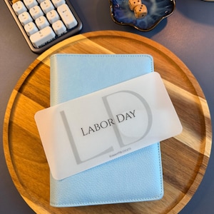 May include: A light blue textured personal planner featuring a translucent rectangular divider card with the text LABOR DAY and the initials LD printed in black, along with the small text KIMMYPBUDGETS at the bottom center of the card.