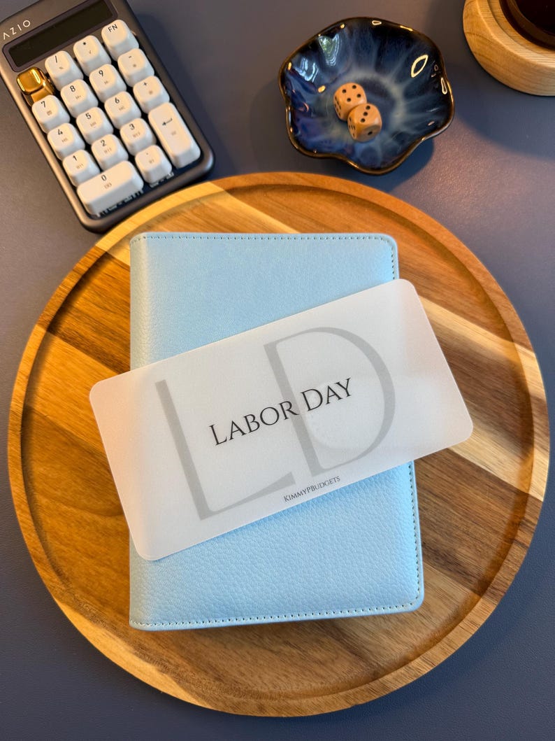 May include: A light blue textured personal planner featuring a translucent rectangular divider card with the text LABOR DAY and the initials LD printed in black, along with the small text KIMMYPBUDGETS at the bottom center of the card.