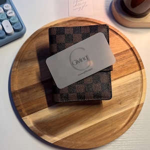 May include: A brown and black checkered wallet featuring a rectangular frosted card with the letter G and the word Giving printed in black. The wallet is positioned on a round wooden tray, with a blue calculator visible in the upper left corner.