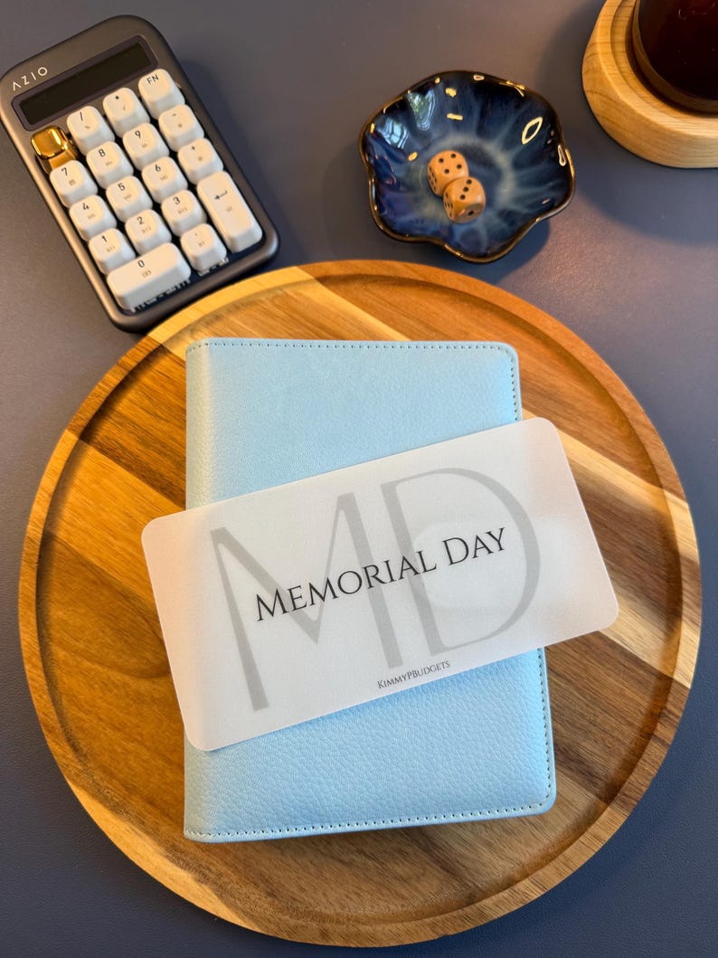 May include: A light blue textured binder featuring a translucent white card overlay with the text MEMORIAL DAY and KIMMYPBUDGETS printed in black. The binder is placed on a round wooden tray alongside a calculator, a blue bowl, and two dice.