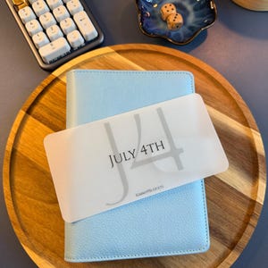 May include: A light blue textured binder featuring a translucent rectangular card overlay with the text JULY 4TH and KIMMYPBUDGETS printed in black. The binder is positioned on a round wooden tray alongside a calculator and a small blue bowl.