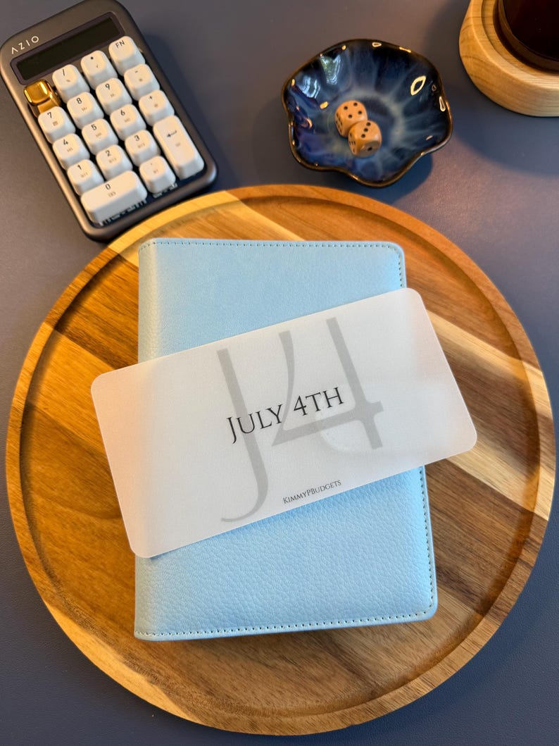 May include: A light blue textured binder featuring a translucent rectangular card overlay with the text JULY 4TH and KIMMYPBUDGETS printed in black. The binder is positioned on a round wooden tray alongside a calculator and a small blue bowl.