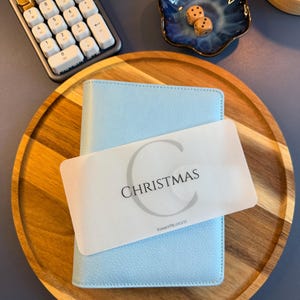 May include: A light blue A6 binder with a textured cover, featuring a translucent white divider card labeled with a large letter C, the word CHRISTMAS, and the text KIMMYPBUDGETS, resting on a round wooden tray with a calculator and dice.