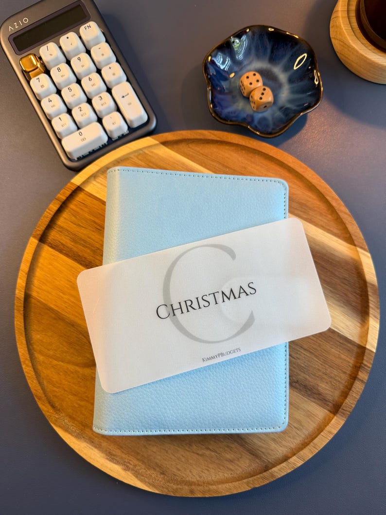 May include: A light blue A6 binder with a textured cover, featuring a translucent white divider card labeled with a large letter C, the word CHRISTMAS, and the text KIMMYPBUDGETS, resting on a round wooden tray with a calculator and dice.
