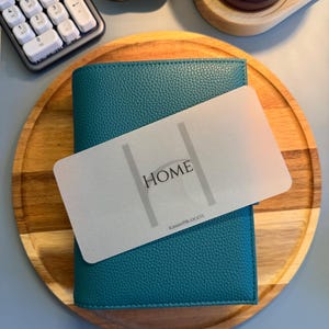 May include: A teal textured leather binder featuring a translucent rectangular divider card placed on top. The divider card displays the word HOME in black capital letters between two vertical lines, with the text KIMMYPBUDGETS printed at the bottom.