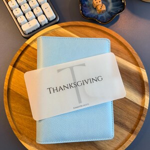 May include: A light blue A6 binder with a textured cover, featuring a translucent white divider card labeled with a large letter T, the word THANKSGIVING, and the text KIMMYPBUDGETS, resting on a round wooden tray with a calculator and dice.