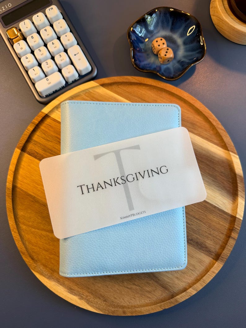 May include: A light blue A6 binder with a textured cover, featuring a translucent white divider card labeled with a large letter T, the word THANKSGIVING, and the text KIMMYPBUDGETS, resting on a round wooden tray with a calculator and dice.
