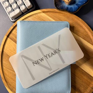 May include: A light blue textured binder with a translucent rectangular card placed on top. The card features the text "NEW YEAR'S" in a bold, dark font, with the letters "NY" overlaid in a larger, lighter gray, and "KIMMYPBUDGETS" at the bottom.