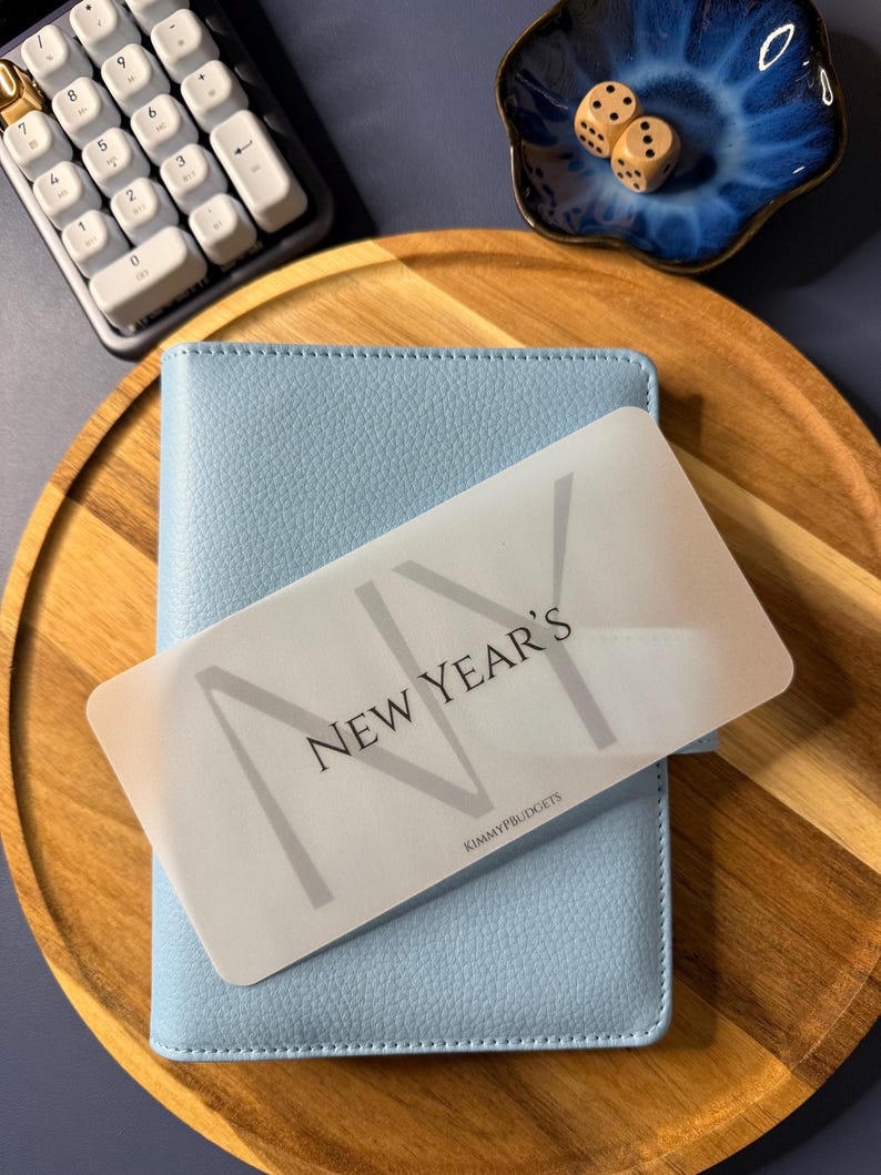 May include: A light blue textured binder with a translucent rectangular card placed on top. The card features the text "NEW YEAR'S" in a bold, dark font, with the letters "NY" overlaid in a larger, lighter gray, and "KIMMYPBUDGETS" at the bottom.