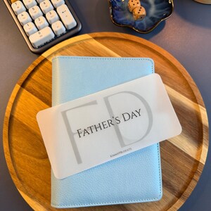 May include: A light blue textured personal planner with a rectangular white card placed on top. The card features the text FATHER'S DAY in black letters with a large grey FD monogram in the background and the small text KIMMYPBUDGETS at the bottom.
