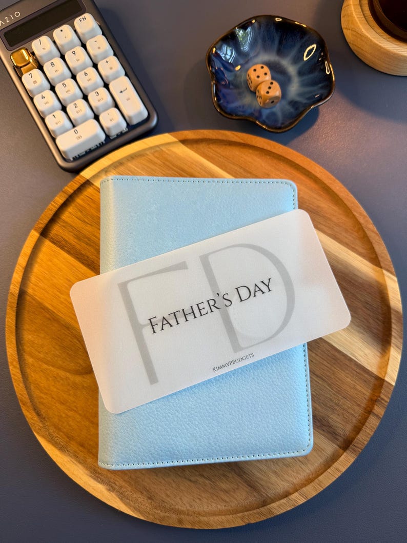 May include: A light blue textured personal planner with a rectangular white card placed on top. The card features the text FATHER'S DAY in black letters with a large grey FD monogram in the background and the small text KIMMYPBUDGETS at the bottom.