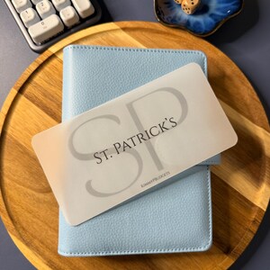 May include: A light blue textured personal planner with a translucent overlay card featuring the text ST. PATRICK'S and KIMMYPBUDGETS in black lettering. The planner is positioned on a round wooden tray alongside a white numeric keypad and a small blue dish containing two wooden dice.