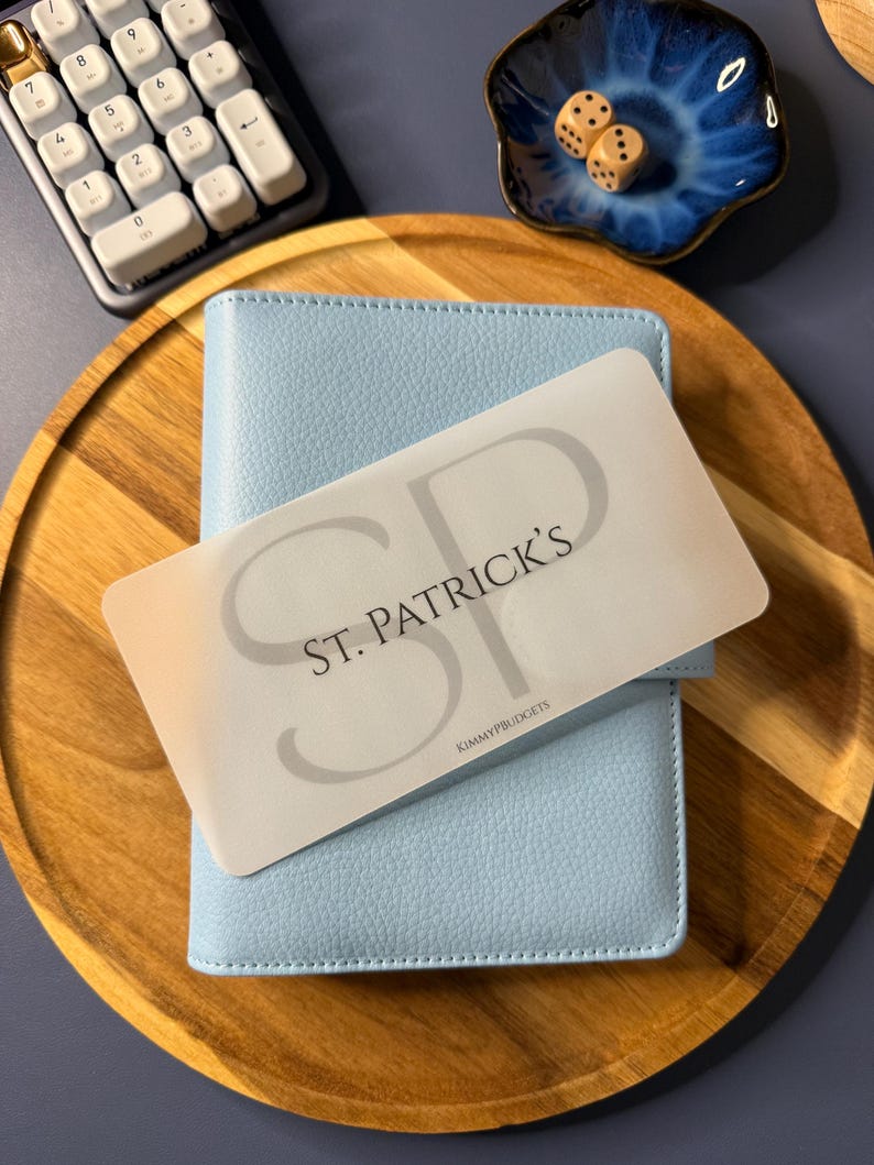 May include: A light blue textured personal planner with a translucent overlay card featuring the text ST. PATRICK'S and KIMMYPBUDGETS in black lettering. The planner is positioned on a round wooden tray alongside a white numeric keypad and a small blue dish containing two wooden dice.