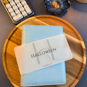 May include: A light blue textured binder with a translucent plastic divider featuring the word HALLOWEEN in black text and the smaller text KIMMYPBUDGETS below it, resting on a round wooden tray alongside a calculator and two dice.