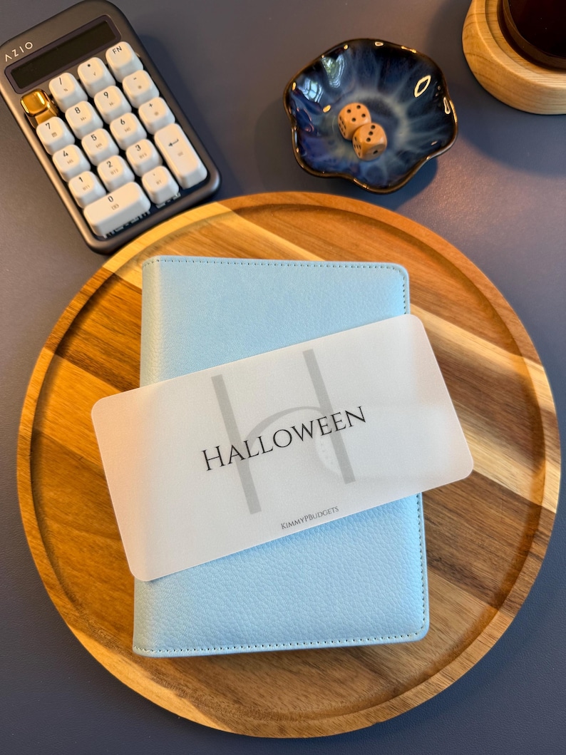 May include: A light blue textured binder with a translucent plastic divider featuring the word HALLOWEEN in black text and the smaller text KIMMYPBUDGETS below it, resting on a round wooden tray alongside a calculator and two dice.