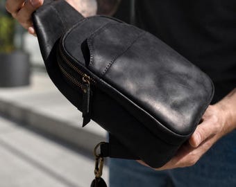 Pode incluir: Uma bolsa tiracolo de couro preto com acabamento fosco liso, um bolso frontal com costura curva e dois compartimentos principais com zíperes de metal escuro. Um gancho giratório de metal está preso à parte inferior da bolsa.
