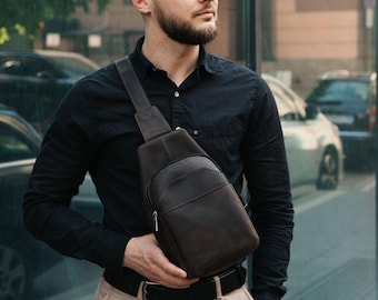 May include: A dark brown leather sling bag with a single shoulder strap. The bag has a vertical design with a zippered pocket and a smooth, polished finish. The bag is worn across the body.