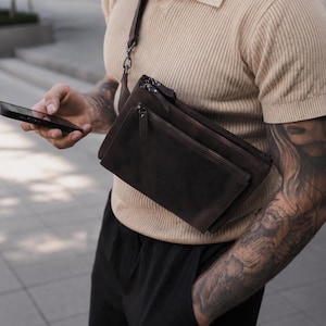 Può includere: Una borsa a tracolla in pelle marrone scuro con silhouette rettangolare, scomparti multipli con cerniera e tracolla regolabile abbinata. La borsa presenta una finitura opaca liscia con cerniere in metallo visibili e tiretti in pelle.