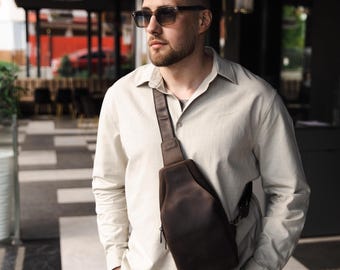 May include: A dark brown leather sling bag with a single shoulder strap and a front zipper. The bag is worn across the body. The person in the image is wearing sunglasses and a light-colored button-down shirt.