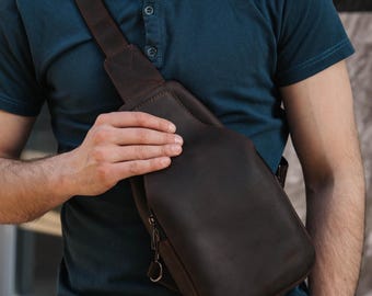 May include: A dark brown leather sling bag with a shoulder strap. The bag has a zippered pocket and a curved design. The person in the image is wearing a dark blue shirt.