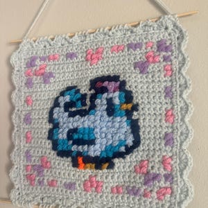 May include: A light blue, square, crocheted wall hanging featuring a pixelated bird design in shades of blue, with pink and purple heart accents. The piece is hung with wooden dowels and a light blue string.