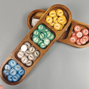 May include: Wooden game token trays filled with colorful game pieces. The tokens feature various designs, including leaves, cherries, and fish. The trays are made of a warm-toned wood and are divided into three sections.
