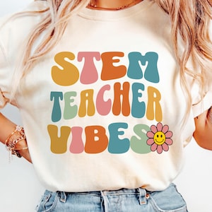 Pode incluir: Camiseta creme com as palavras "STEM TEACHER VIBES" em letras coloridas em estilo retrô. Um gráfico de uma flor sorridente está ao lado da palavra "VIBES". É uma peça casual para o dia a dia.