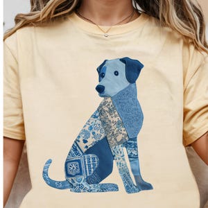 May include: A light yellow t-shirt featuring a seated dog design. The dog is composed of various blue fabric patterns, creating a patchwork effect. The dog's head and ears are a solid blue, and the overall design is centered on the shirt.