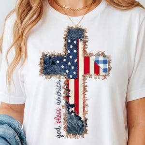 Puede incluir: Camiseta blanca con un diseño de cruz con temática de la bandera estadounidense. La cruz está hecha de parches de mezclilla y tela, con las palabras "god bless america" en una fuente cursiva.