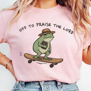 Puede incluir: Camiseta rosa claro con un dibujo de una rana en monopatín, con sombrero y libro. El texto "OFF TO PRAISE THE LORD" está impreso en negro sobre la rana.