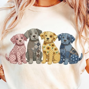 May include: Cream-colored sweatshirt featuring four cartoon puppies. Each puppy has a unique patchwork design: pink hearts, gray argyle, yellow sunflowers, and blue stars. The puppies are sitting, with their ears perked up.