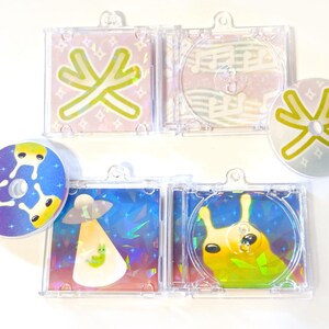 May include: A collection of colorful CDs and CD cases with whimsical designs. The designs include an alien, a snail, and abstract shapes in various colors. The cases are clear plastic, and the CDs are round.