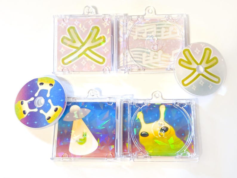 May include: A collection of colorful CDs and CD cases with whimsical designs. The designs include an alien, a snail, and abstract shapes in various colors. The cases are clear plastic, and the CDs are round.