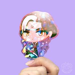 May include: A holographic sticker of a chibi character with white hair and a determined expression. The sticker has a purple and iridescent finish, with diamond-shaped accents on the character's clothing. The sticker is held against a light purple background.
